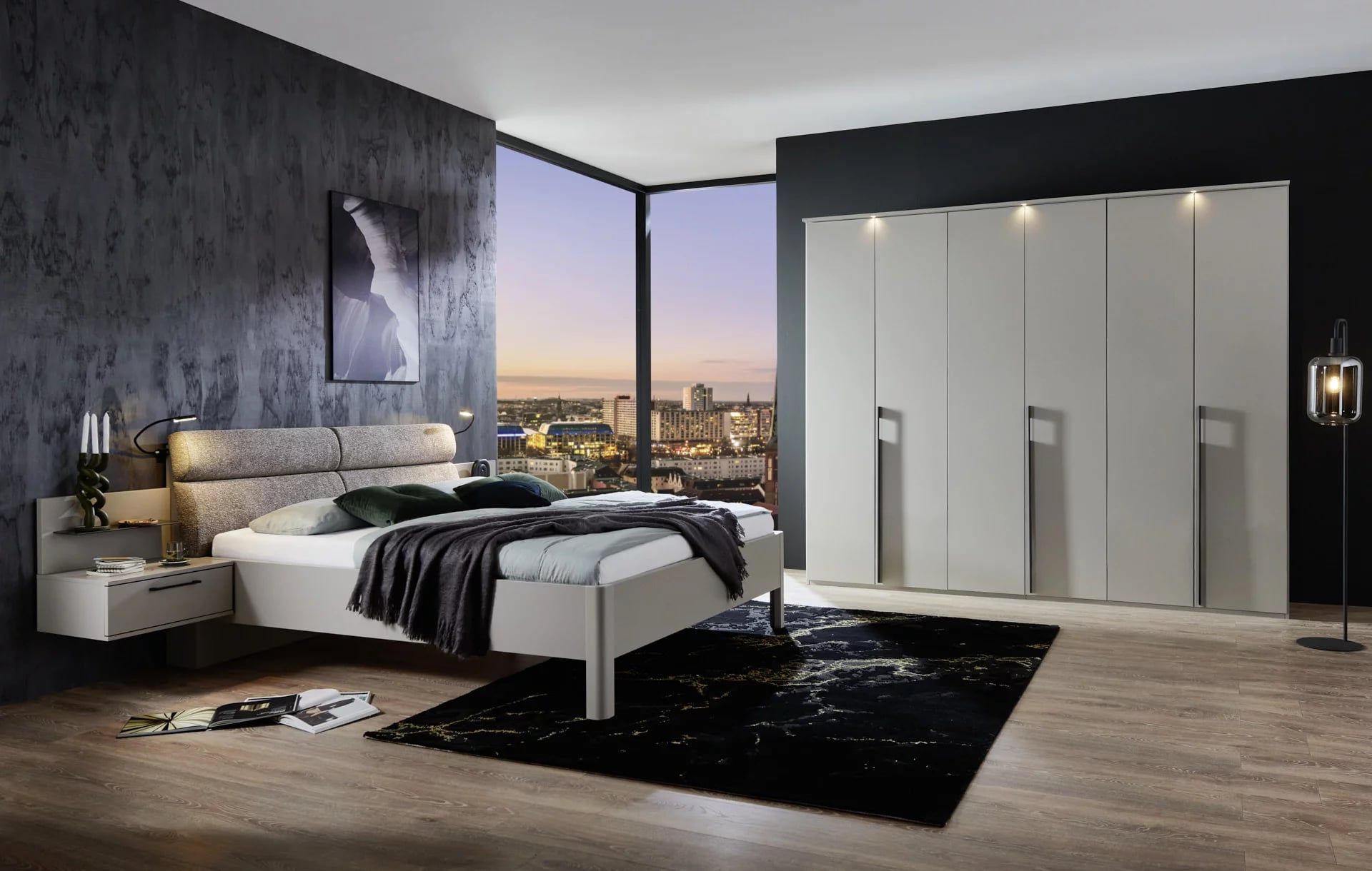 Schlafzimmer mit Polsterkopfteil, Nachttischen und hellem Drehtürenschrank vor großer Fensterfront mit Blick auf die Stadt