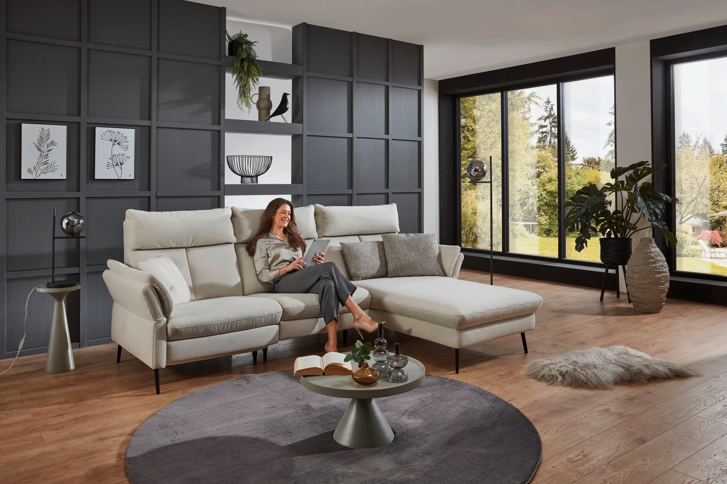 Ecksofa in Creme mit Kopfstützen und Longchair in einem Wohnzimmer mit dunkler Regalwand und großen Fenstern